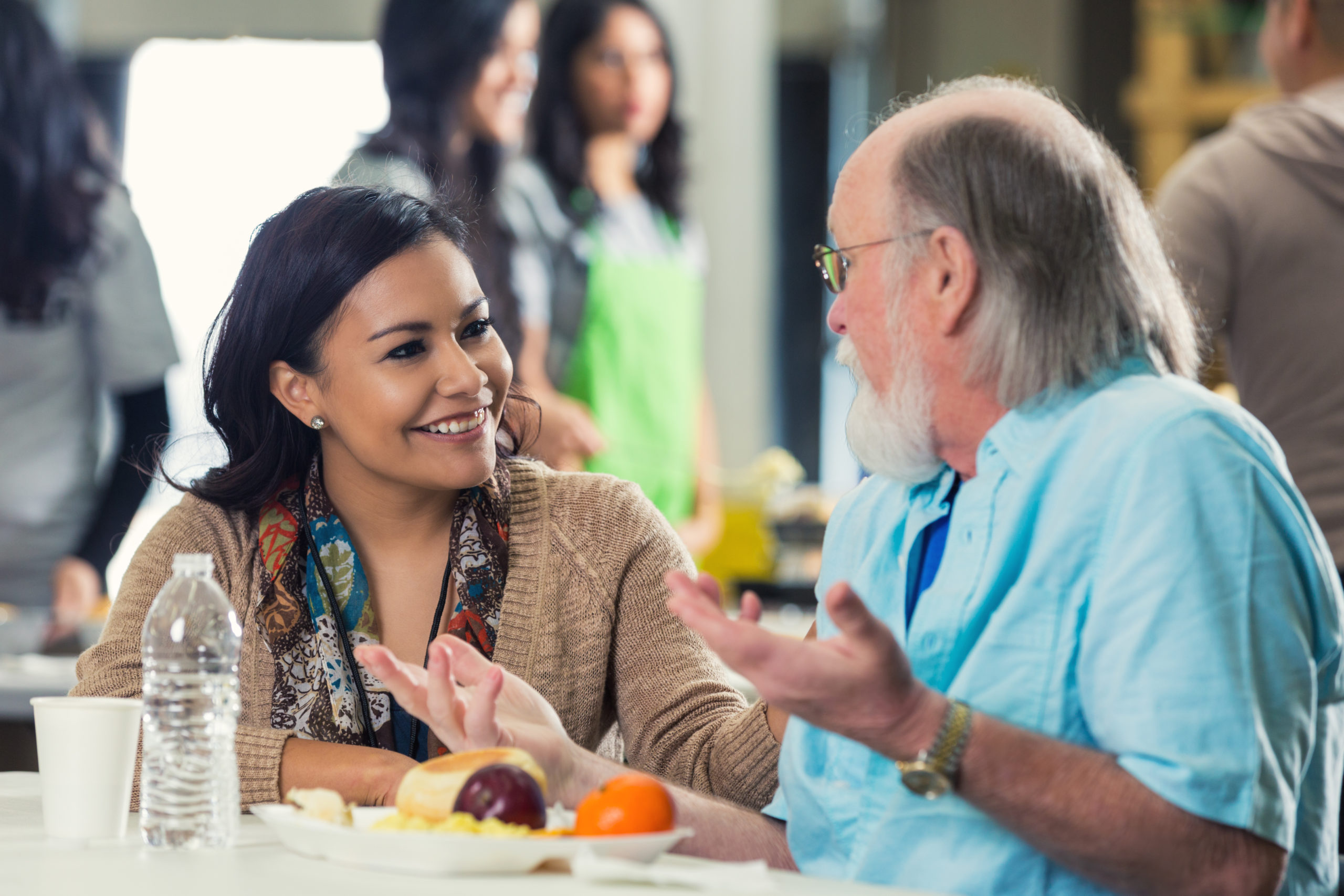 Young adult Hispanic woman is sitting at table in soup kitchen and talking with senior Caucasian man. Man has grey hair and beard. They are discussing something while senior enjoys healthy meal. Volunteers are serving food in background.