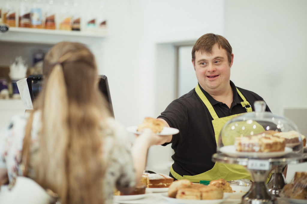 A man with Down syndrome is serving a scone to a woman in a cafe where he works.