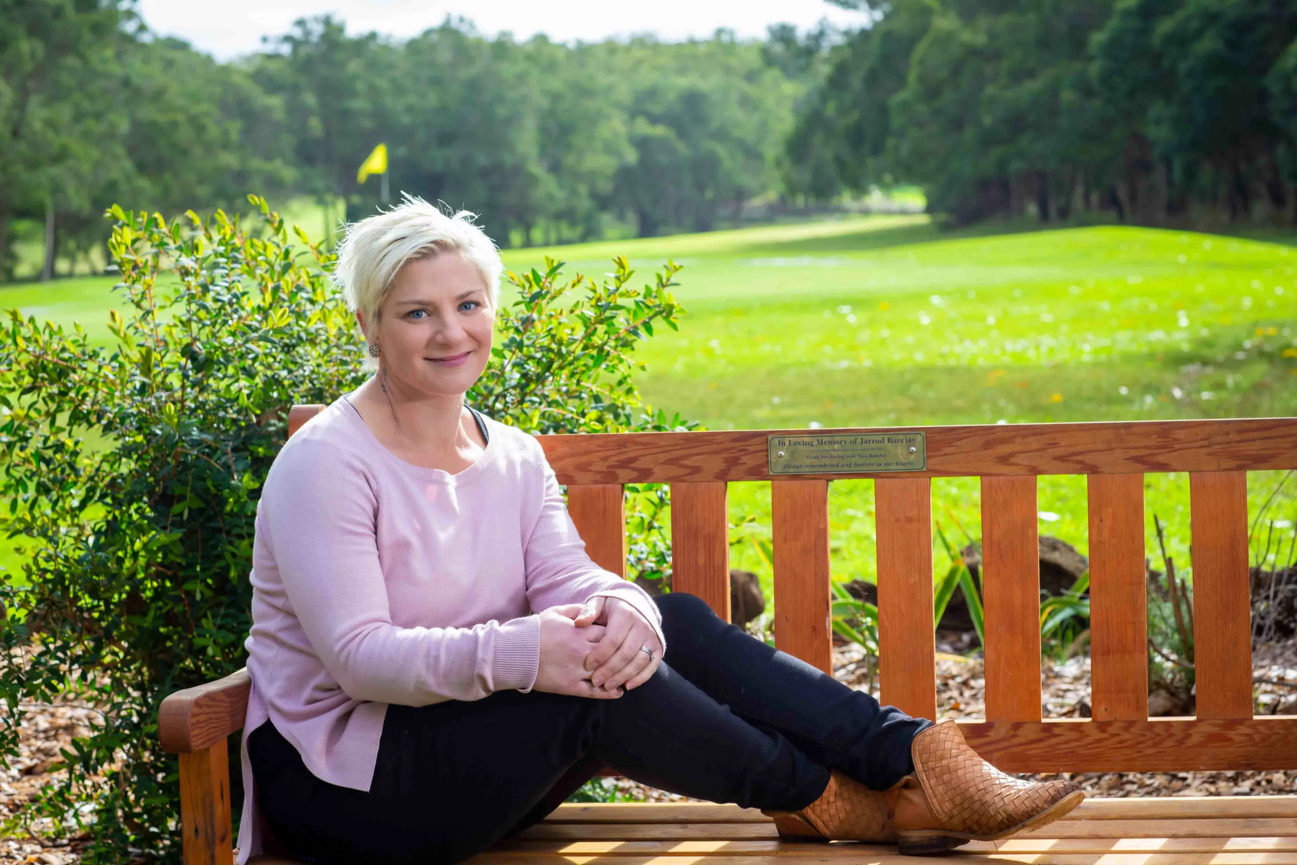 Nea Barclay is seated on the wooden seat in memory of her husband at the Heywood Golf Club in Victoria. Nea wears a lilac jumper, dark pants and tan boots. A garden bed and golf green with a yellow flag are behind her.