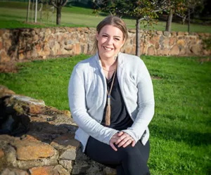 Melissa is smiling and seated outside on a semi circular stone wall surrounded by green grass. She is wearing black pans and top and a grey jacket. 